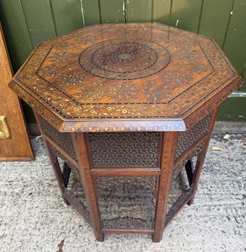 An octagonal Anglo-Indian teak side table featuring intricate floral and geometric brass and copper wire inlay on its top surface, set upon a carved and pierced folding lattice wooden base.