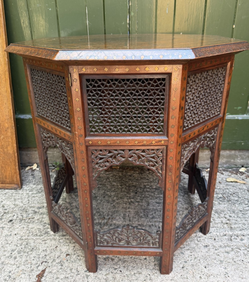 An octagonal Anglo-Indian teak side table featuring intricate floral and geometric brass and copper wire inlay on its top surface, set upon a carved and pierced folding lattice wooden base. View II