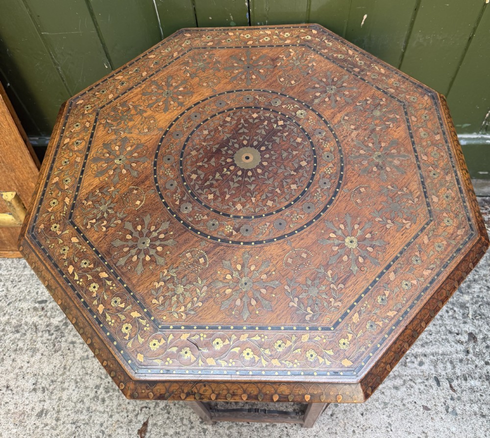 An octagonal Anglo-Indian teak side table featuring intricate floral and geometric brass and copper wire inlay on its top surface, set upon a carved and pierced folding lattice wooden base. View XX