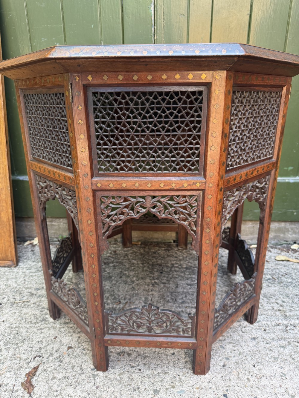 An octagonal Anglo-Indian teak side table featuring intricate floral and geometric brass and copper wire inlay on its top surface, set upon a carved and pierced folding lattice wooden base. View XXI