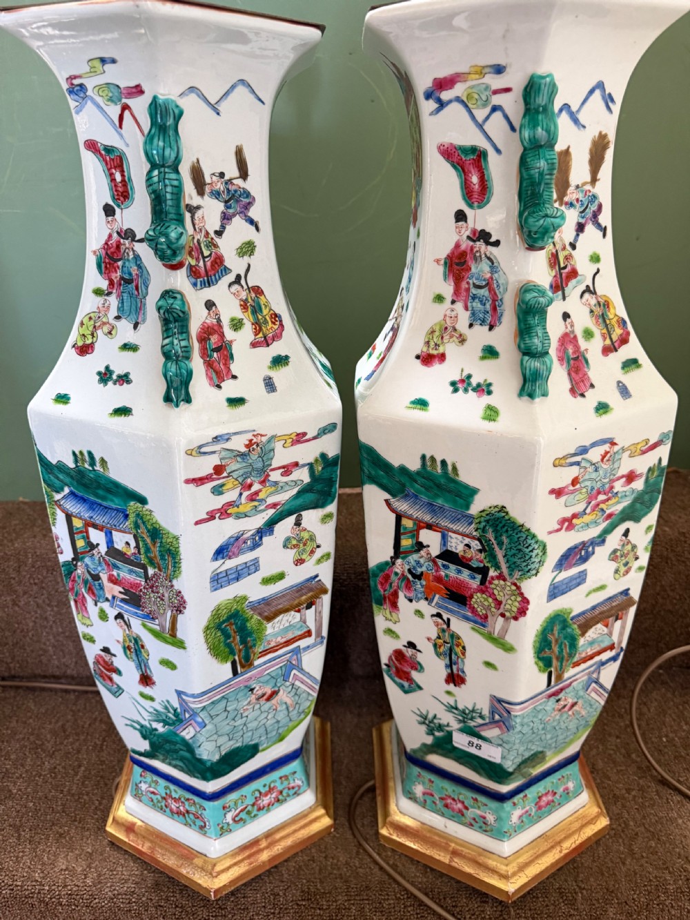 A pair of hexagonal Chinese famille-verte porcelain lamps decorated with figures in traditional scenes and set on gilded bases. View V