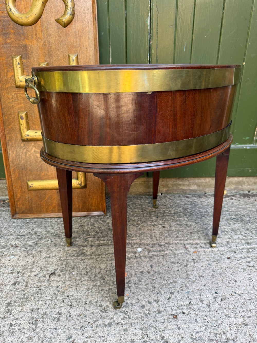 An 18th-century George III mahogany oval wine cooler with thick brass bands and brass handles, resting on a four-legged tapered stand with castors. View III
