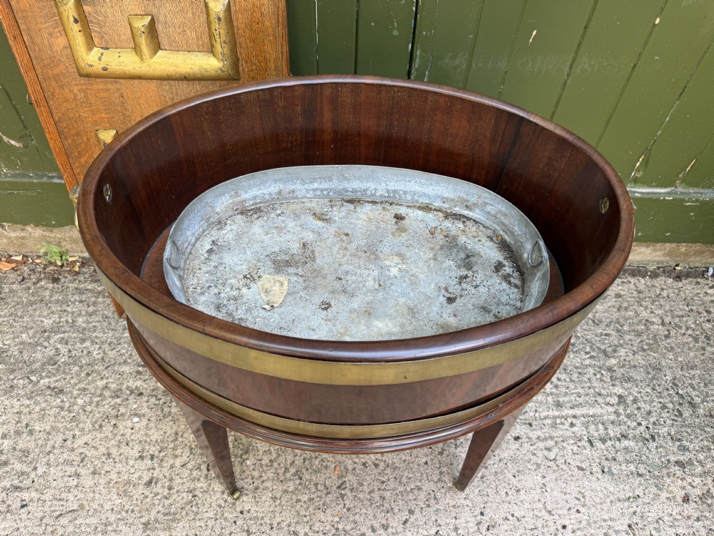 An 18th-century George III mahogany oval wine cooler with thick brass bands and brass handles, resting on a four-legged tapered stand with castors. View VI