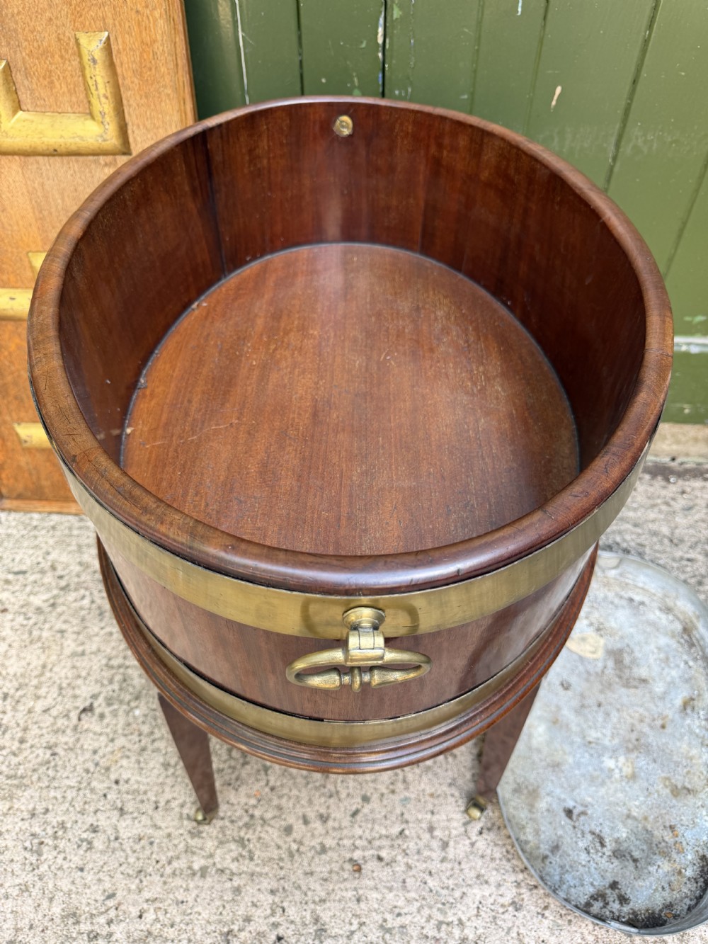 An 18th-century George III mahogany oval wine cooler with thick brass bands and brass handles, resting on a four-legged tapered stand with castors. View X