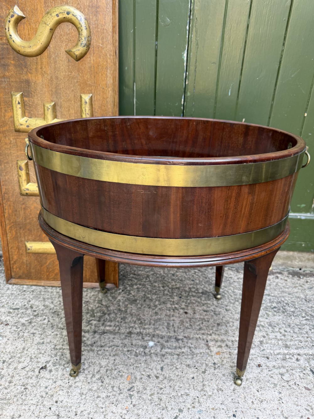 An 18th-century George III mahogany oval wine cooler with thick brass bands and brass handles, resting on a four-legged tapered stand with castors. View XV