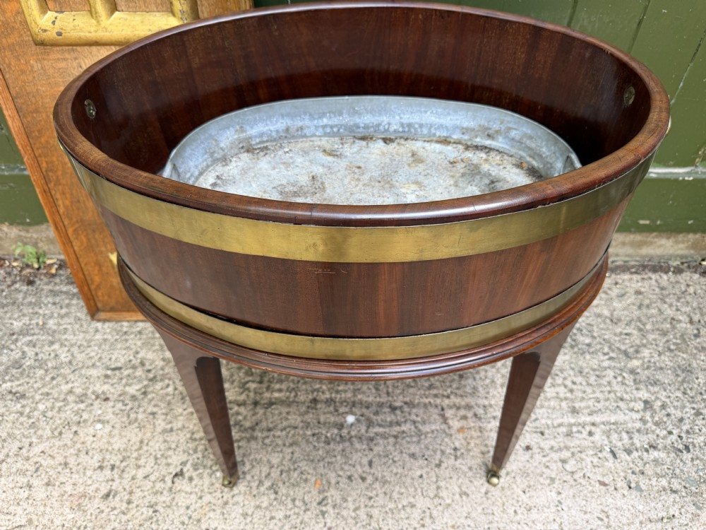 An 18th-century George III mahogany oval wine cooler with thick brass bands and brass handles, resting on a four-legged tapered stand with castors. View XVIII
