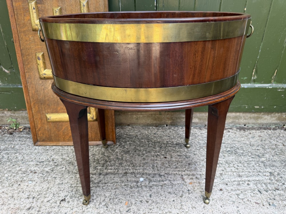 An 18th-century George III mahogany oval wine cooler with thick brass bands and brass handles, resting on a four-legged tapered stand with castors. View XIX