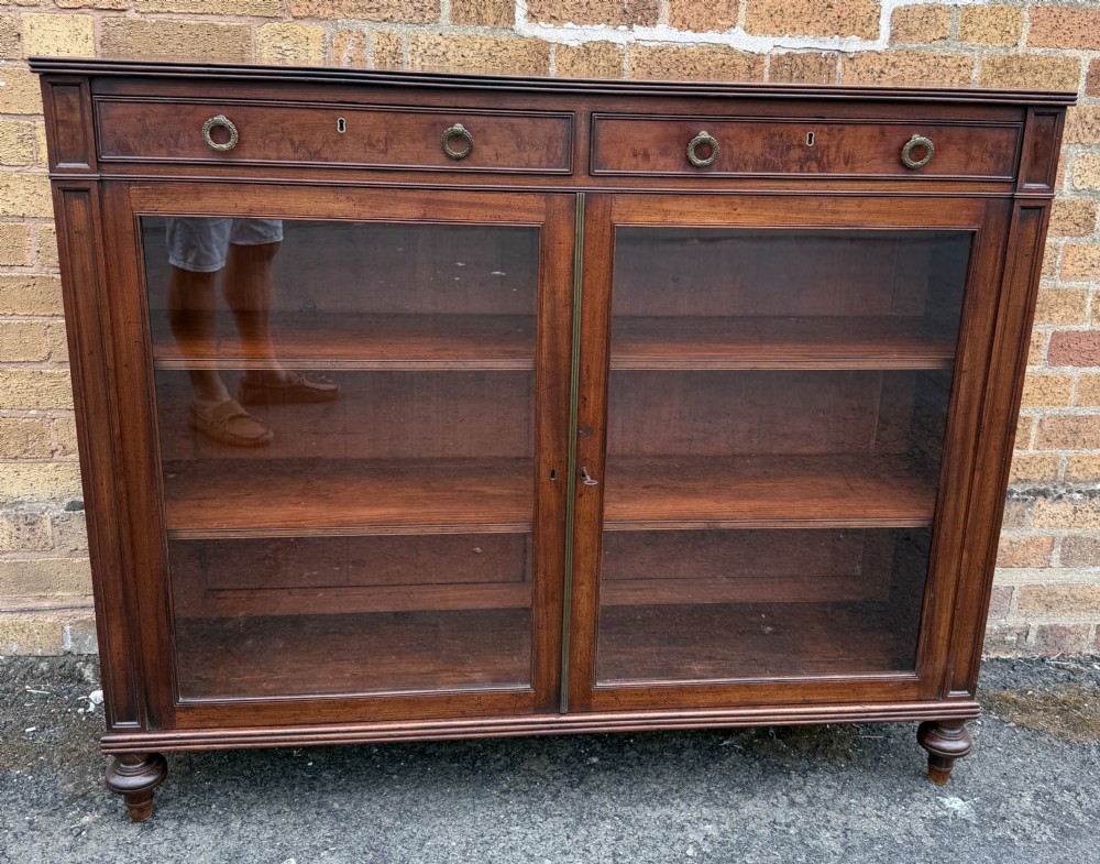 An early nineteenth-century George III mahogany low bookcase featuring two top frieze drawers over glazed twin doors and turned feet. View XX