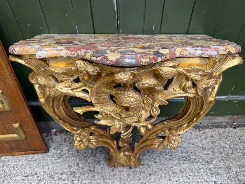 An 18th-century style French carved giltwood wall console table featuring intricate foliage, a central bird motif, and a variegated pink and ochre Breche dAlep marble top. View VI