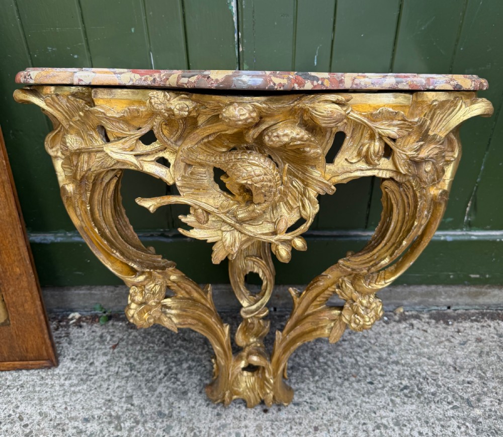 An 18th-century style French carved giltwood wall console table featuring intricate foliage, a central bird motif, and a variegated pink and ochre Breche dAlep marble top. View IX