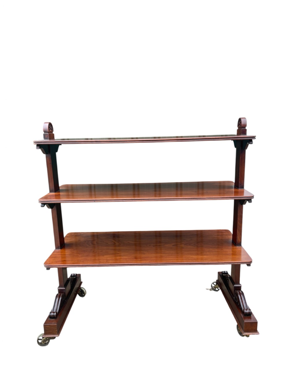 A nineteenth-century mahogany three-tiered dumb waiter featuring carved side uprights and fitted with brass castors at the base.