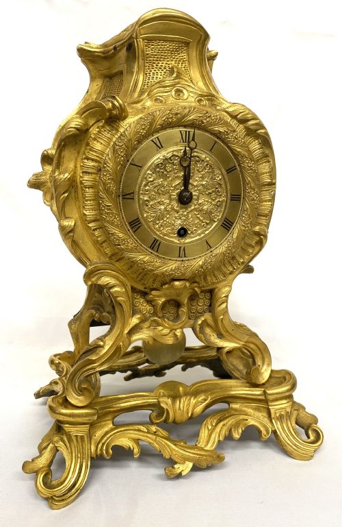 fig 1. A Rococo style gilt bronze mantel clock featuring scrollwork, acanthus leaf carving, and a gilded dial with Roman numerals.