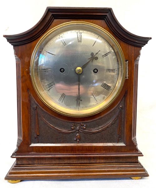fig 1. An antique mahogany double chain fusee mantel clock featuring a curved pediment, fluted side columns, carved drapery swag panel, and a silvered dial with Roman numerals.