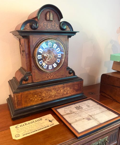 fig 1. An antique walnut and ebonised cuckoo mantel clock featuring floral marquetry inlays, an architectural domed pediment, and an enamel cartouche dial.