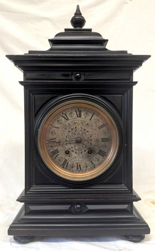 fig 1. An ebonised wooden mantel clock featuring a stepped pedestal base, turned finial, and an engraved silvered metal dial with Roman numerals.
