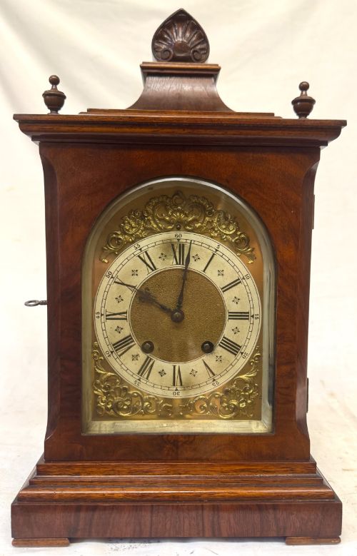 fig 1. An antique walnut bracket clock featuring an arched brass dial with decorative corner spandrels, Roman numerals, and turned finials on a stepped wood base.