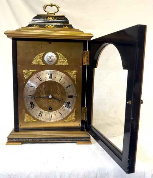 fig 2. A black lacquered chinoiserie bracket clock featuring hand-painted gilt landscape scenes, a brass dial with Roman numerals, and a top brass carrying handle.