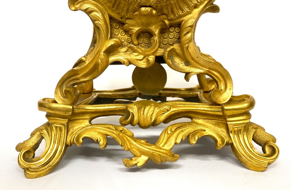 A Rococo style gilt bronze mantel clock featuring scrollwork, acanthus leaf carving, and a gilded dial with Roman numerals. View V