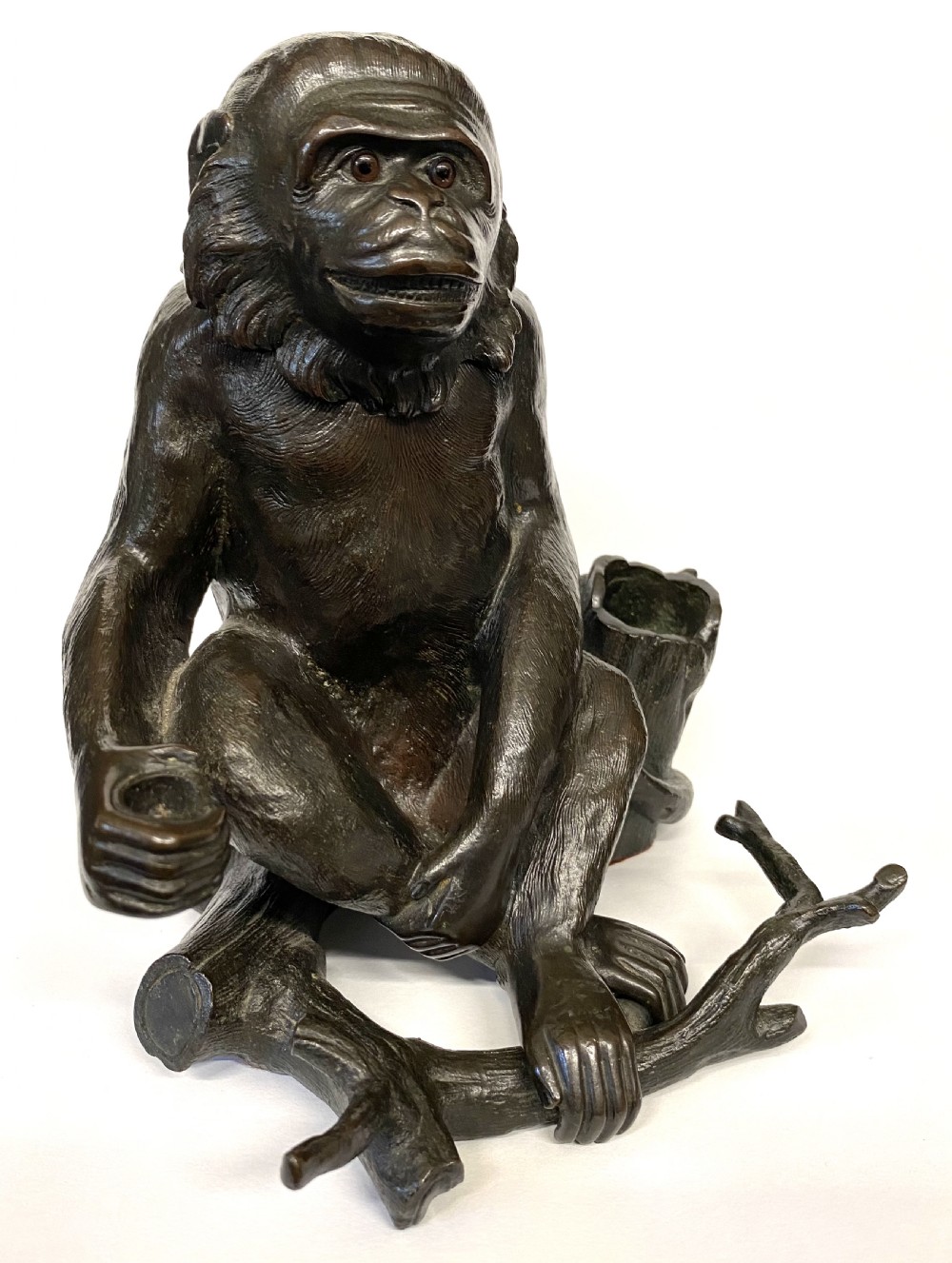 A dark patinated bronze inkwell sculpted in the form of a seated monkey perched on a tree branch with inset glass eyes.
