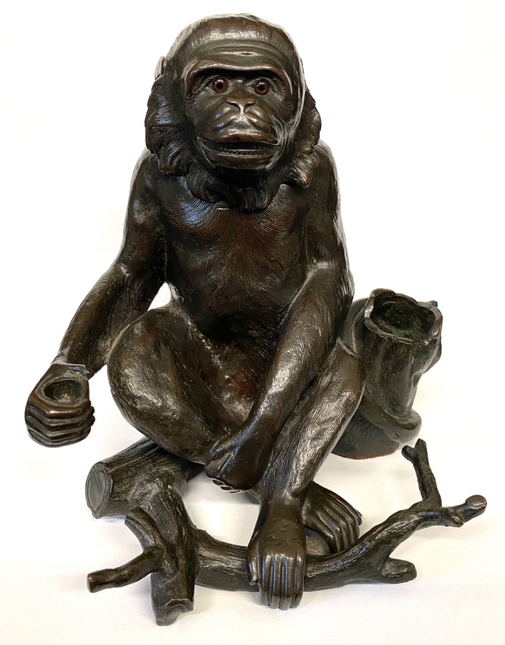 A dark patinated bronze inkwell sculpted in the form of a seated monkey perched on a tree branch with inset glass eyes. View IV