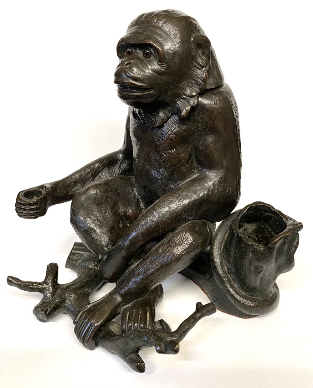 A dark patinated bronze inkwell sculpted in the form of a seated monkey perched on a tree branch with inset glass eyes. View V