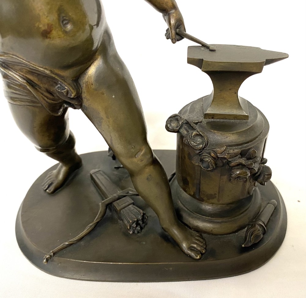 A dark patinated bronze statuette depicting Cupid raising a hammer to forge an arrow on an anvil, resting on a moulded oval base. View V