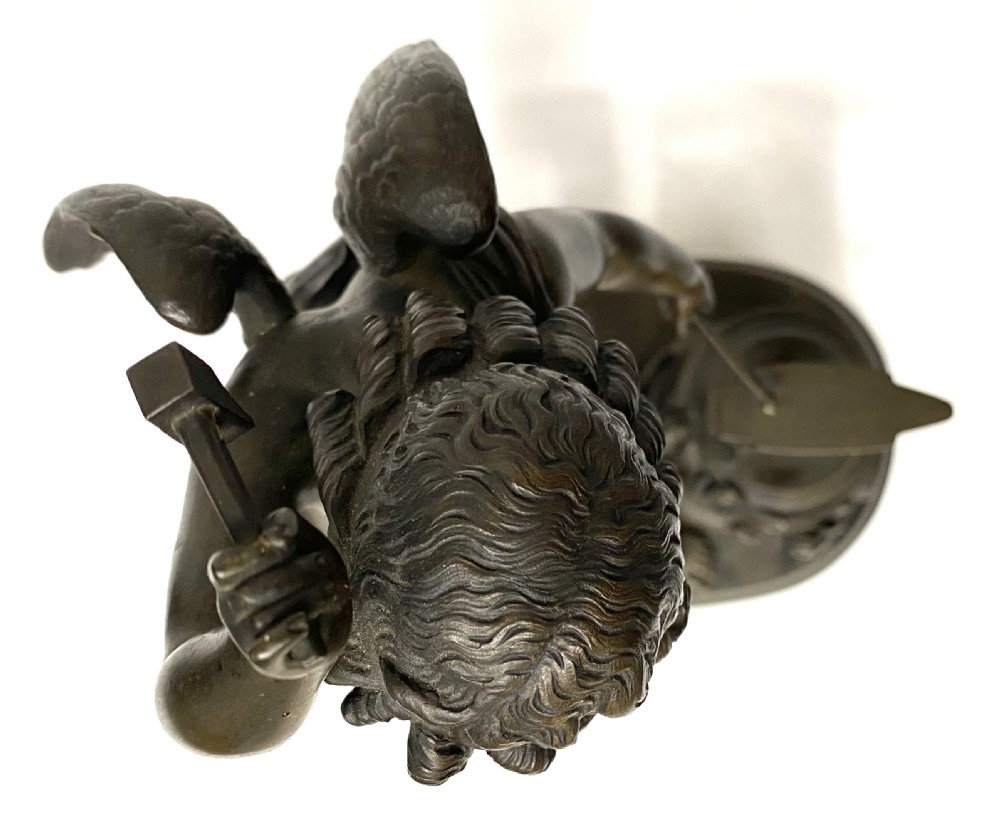 A dark patinated bronze statuette depicting Cupid raising a hammer to forge an arrow on an anvil, resting on a moulded oval base. View XIII