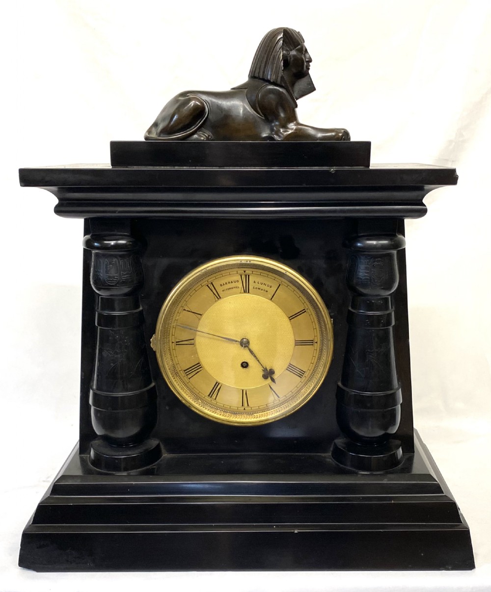 An antique black slate mantel clock in the Egyptian Revival style, featuring a bronze sphinx atop the case and engraved columns framing a circular brass dial.