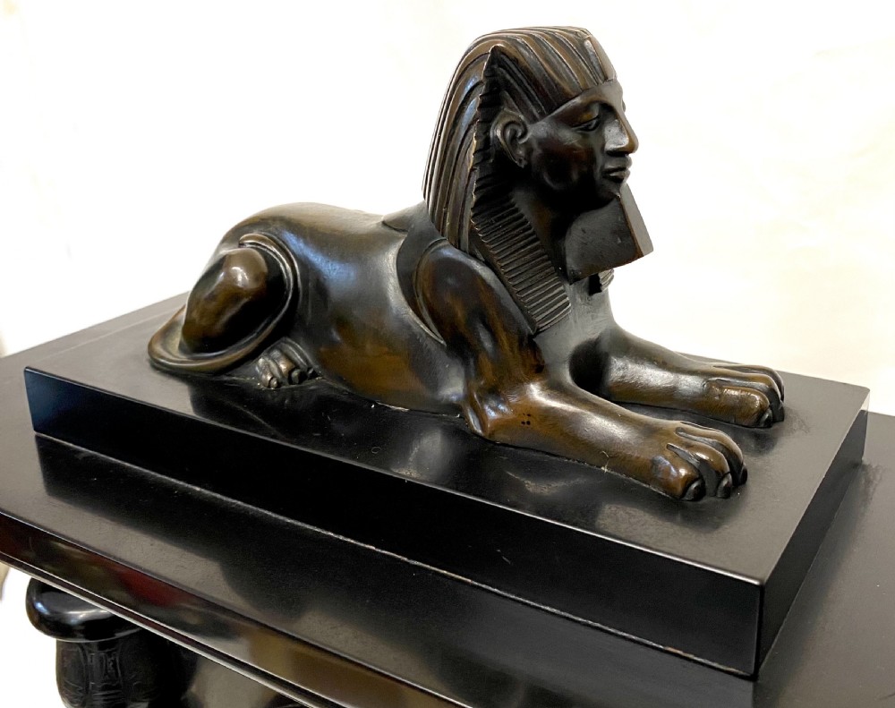 An antique black slate mantel clock in the Egyptian Revival style, featuring a bronze sphinx atop the case and engraved columns framing a circular brass dial. View V