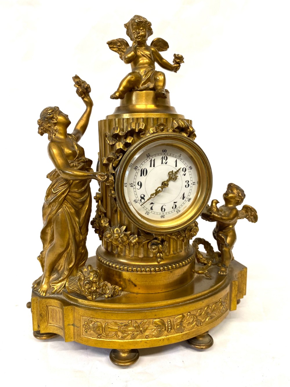 An ornate gilt bronze miniature mantel clock featuring figures of a classical maiden and two winged cherubs surrounding a white enamel dial.