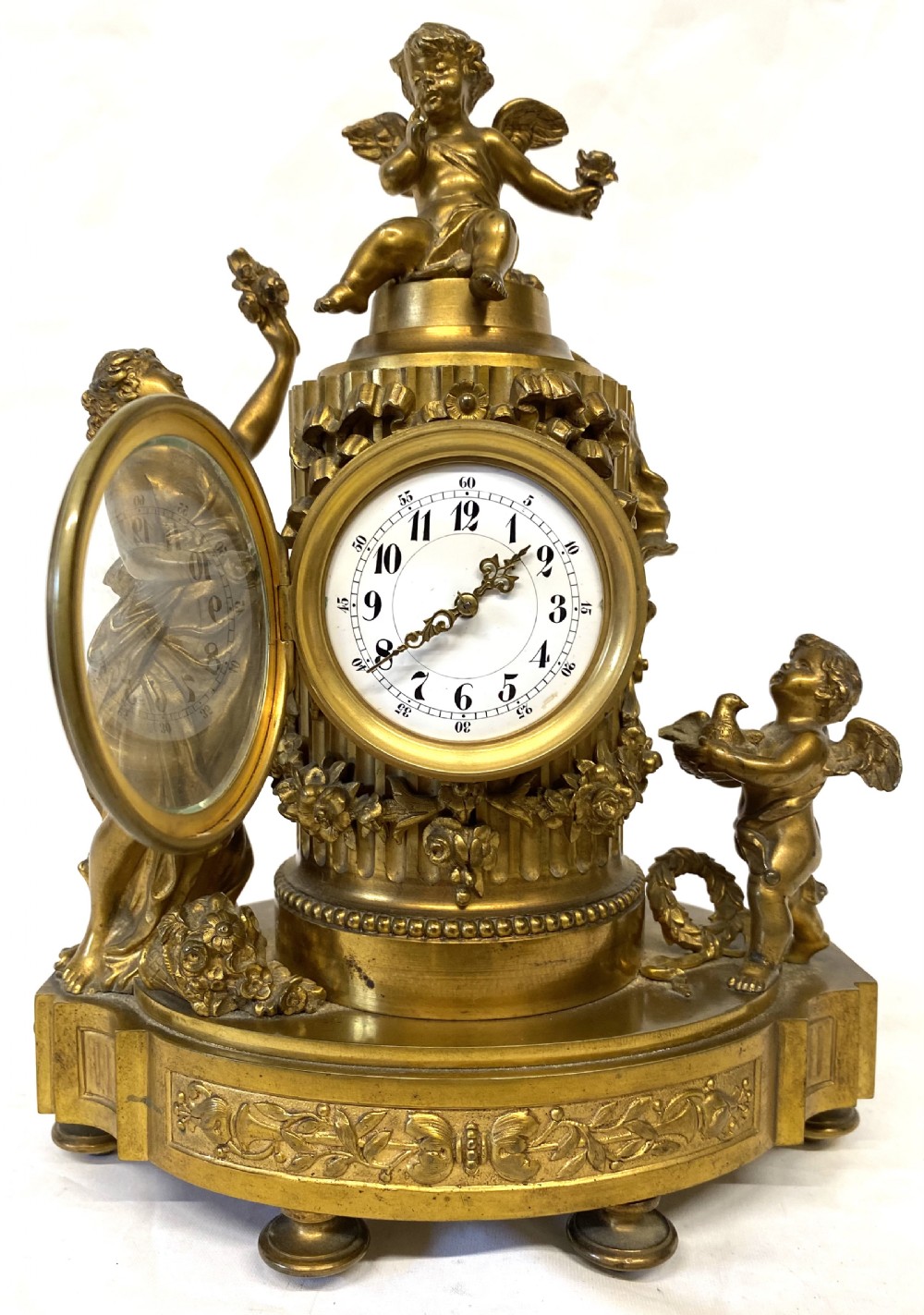 An ornate gilt bronze miniature mantel clock featuring figures of a classical maiden and two winged cherubs surrounding a white enamel dial. View VI
