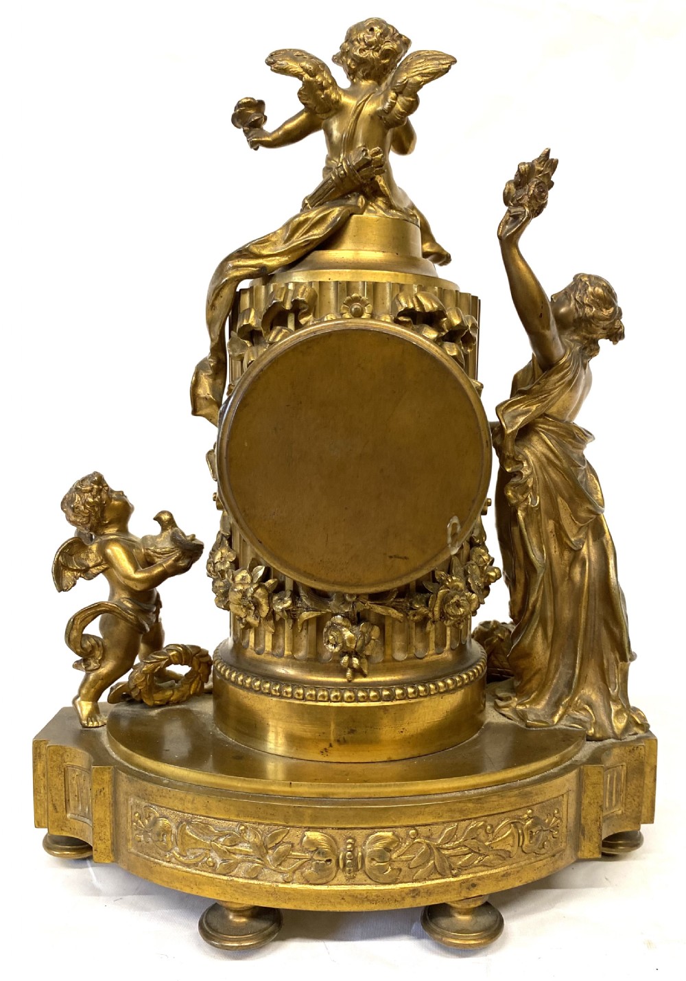 An ornate gilt bronze miniature mantel clock featuring figures of a classical maiden and two winged cherubs surrounding a white enamel dial. View IX