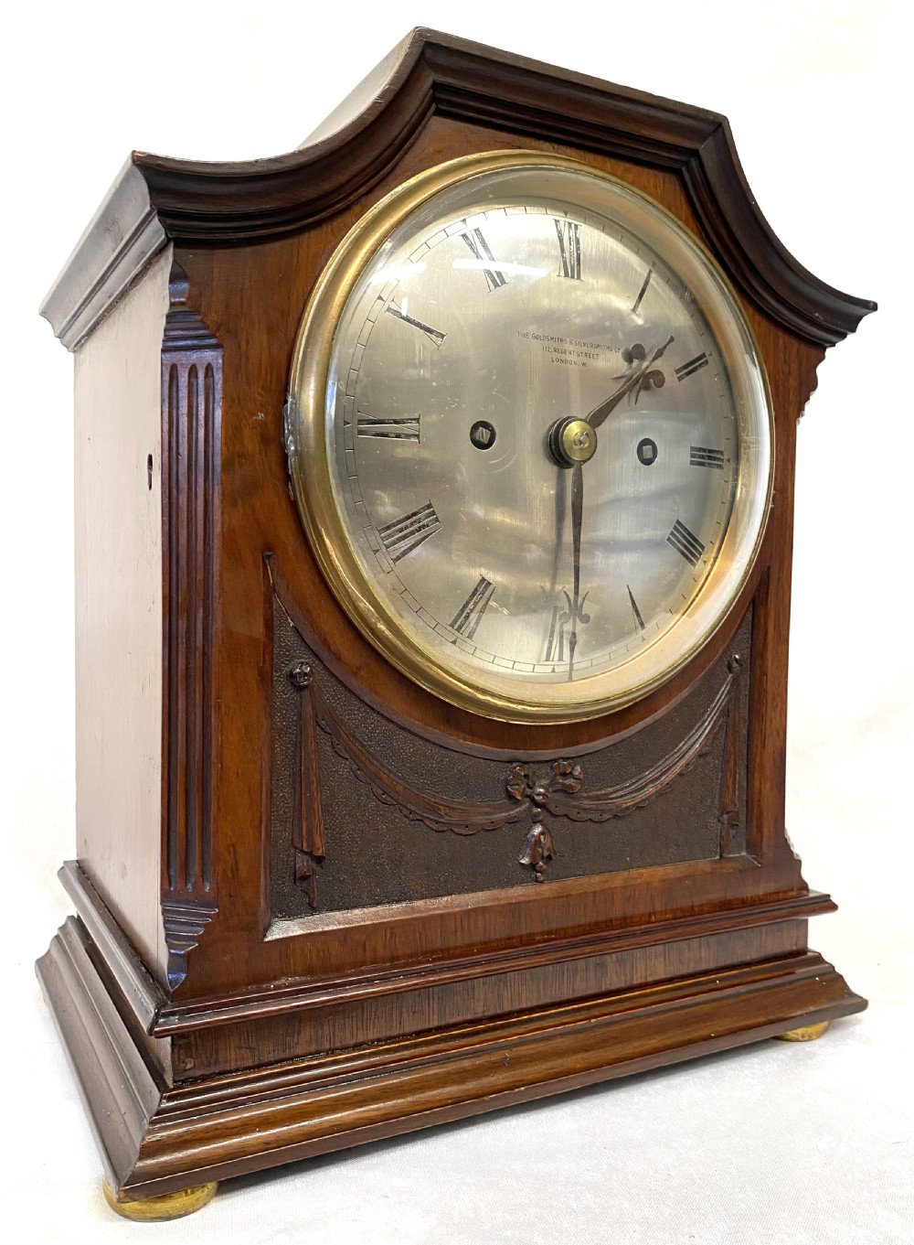 An antique mahogany double chain fusee mantel clock featuring a curved pediment, fluted side columns, carved drapery swag panel, and a silvered dial with Roman numerals.