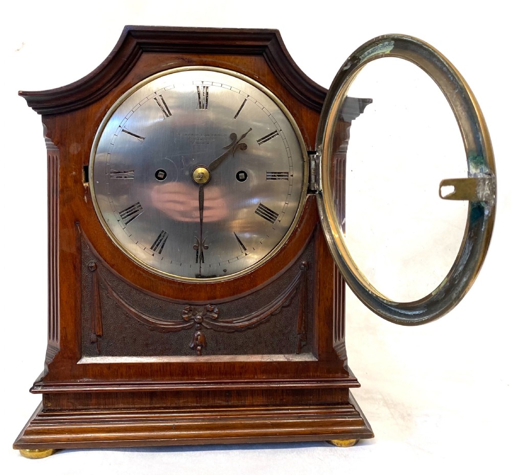 An antique mahogany double chain fusee mantel clock featuring a curved pediment, fluted side columns, carved drapery swag panel, and a silvered dial with Roman numerals. View III