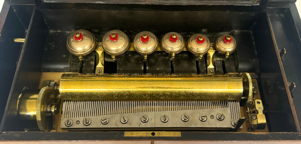 An antique rectangular wooden music box featuring floral marquetry inlay on the lid and front, framed by dark ebonised edges with a key inserted in the lock. View IX