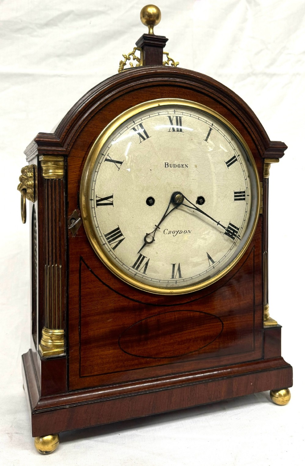 An antique mahogany bracket clock featuring an arched top with a brass finial, fluted brass side columns, and a white dial signed Budgen Croydon.