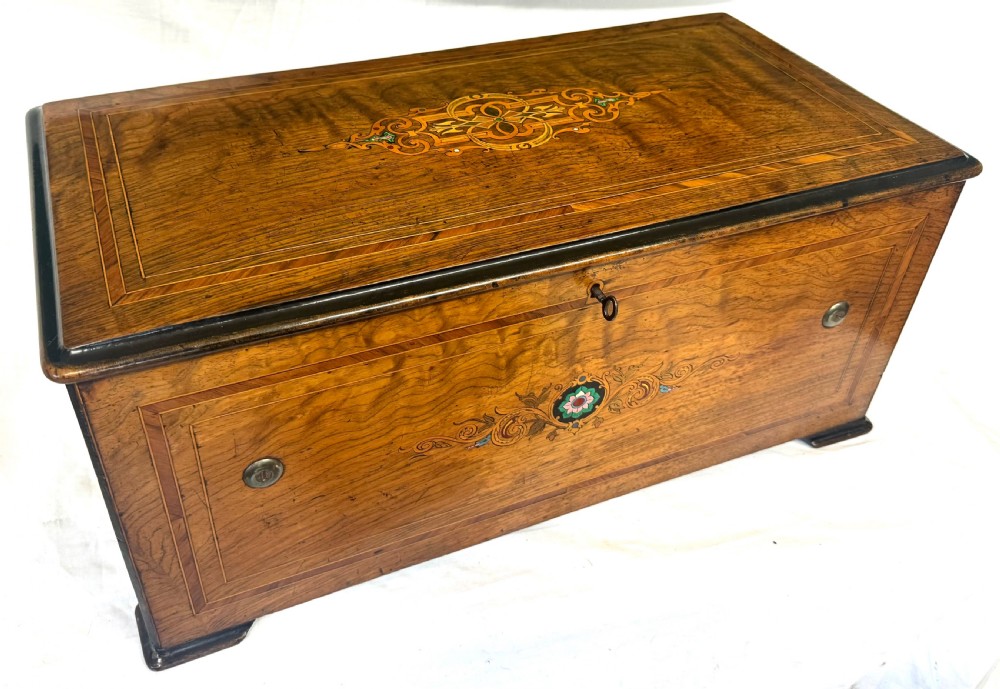 A rectangular rosewood music box featuring floral marquetry inlays on the lid and front panel, flanked by ebonised borders with a key inserted.