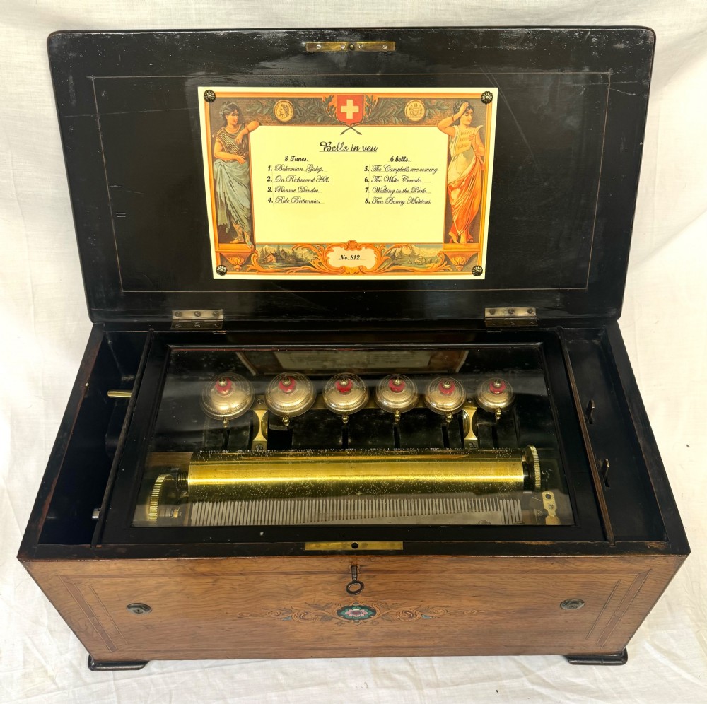 A rectangular rosewood music box featuring floral marquetry inlays on the lid and front panel, flanked by ebonised borders with a key inserted. View VI