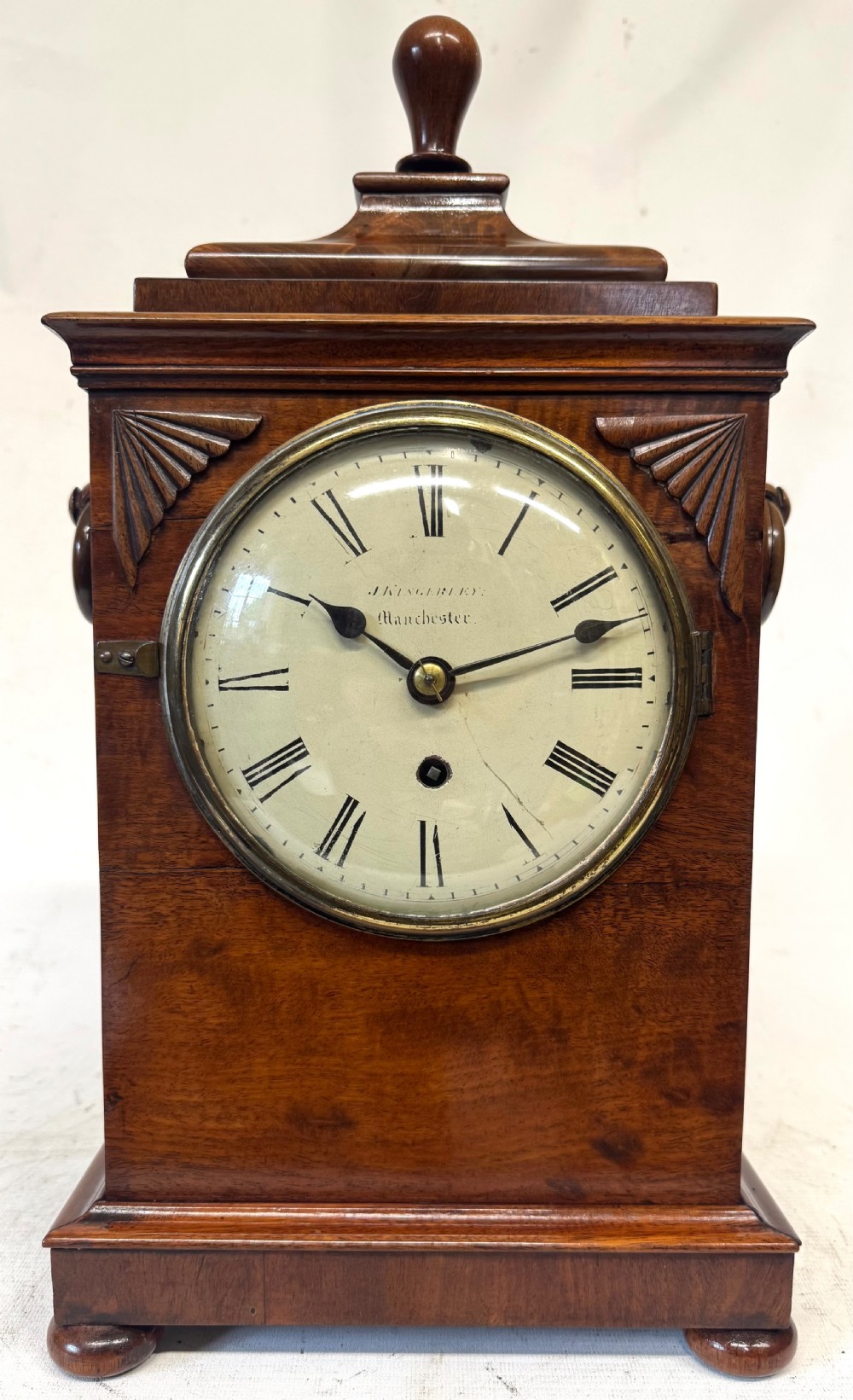 A mahogany single fusee mantel clock featuring a turned finial top, carved fan spandrels, a white dial with Roman numerals signed J Kingerley Manchester, and bun feet.