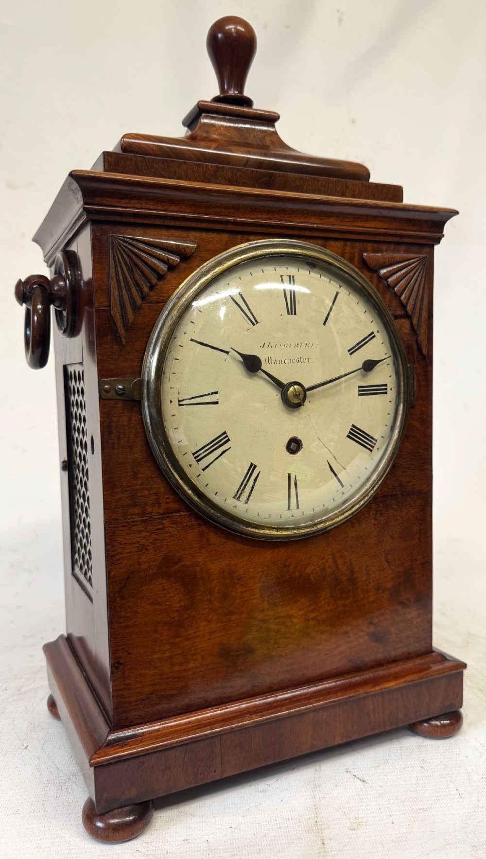 A mahogany single fusee mantel clock featuring a turned finial top, carved fan spandrels, a white dial with Roman numerals signed J Kingerley Manchester, and bun feet. View II