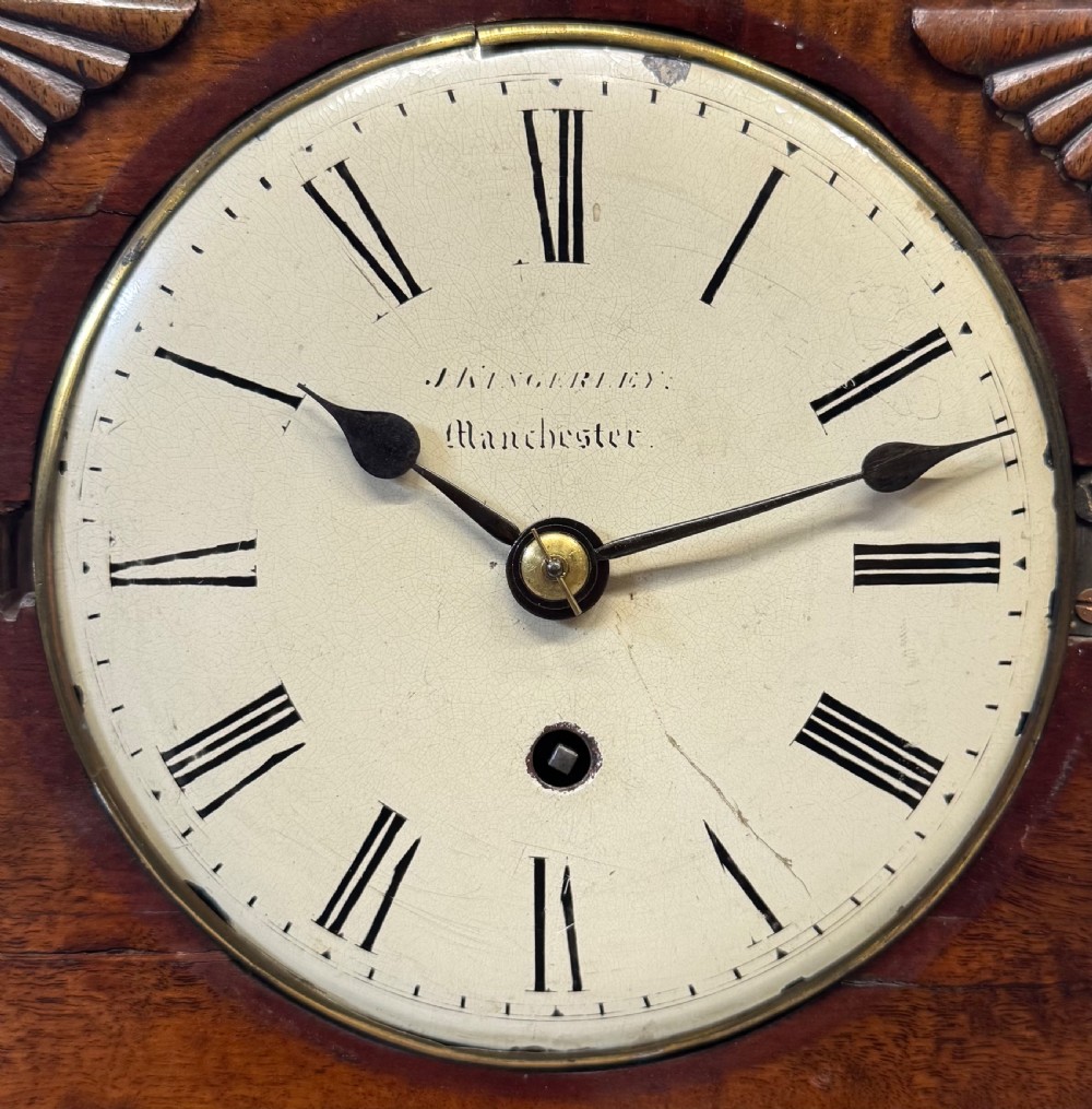 A mahogany single fusee mantel clock featuring a turned finial top, carved fan spandrels, a white dial with Roman numerals signed J Kingerley Manchester, and bun feet. View IV