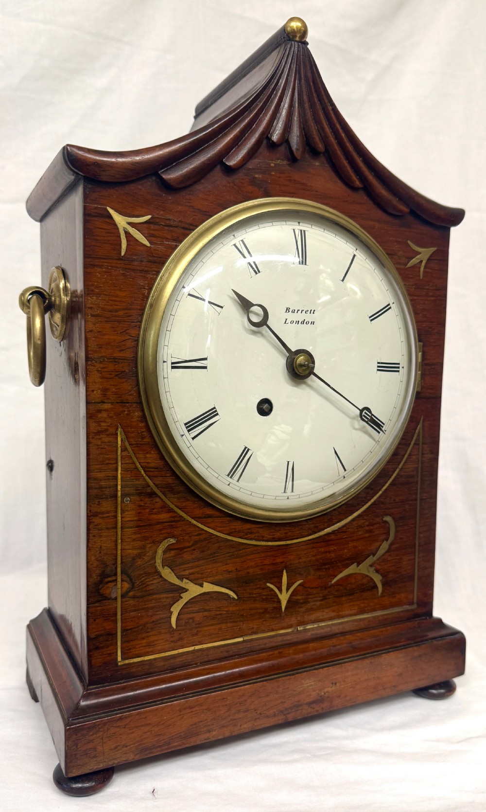 An antique mahogany mantel clock featuring a carved pagoda top, brass inlay details, side ring handles, and a white circular dial signed Barrett London with Roman numerals.