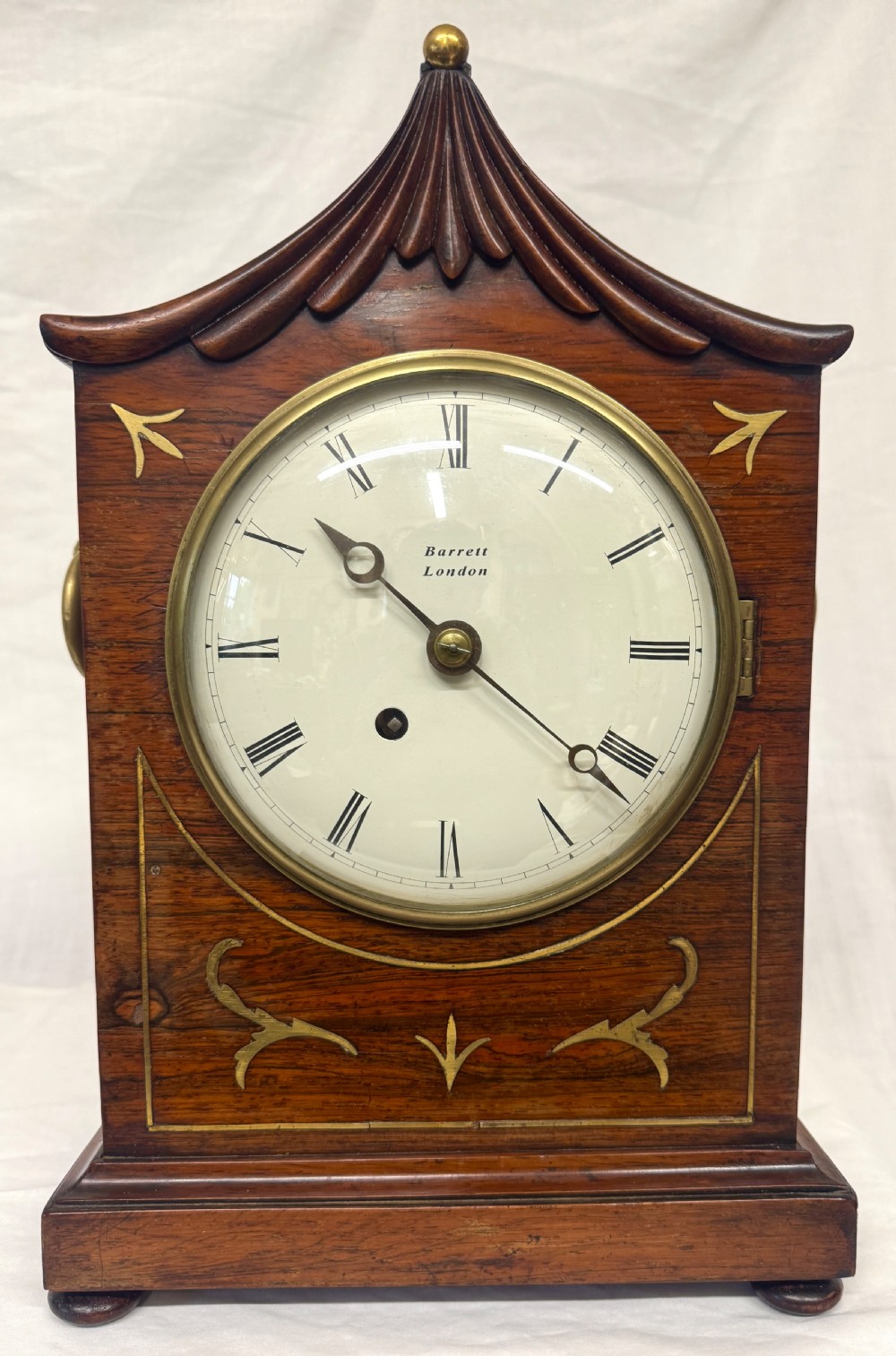 An antique mahogany mantel clock featuring a carved pagoda top, brass inlay details, side ring handles, and a white circular dial signed Barrett London with Roman numerals. View II