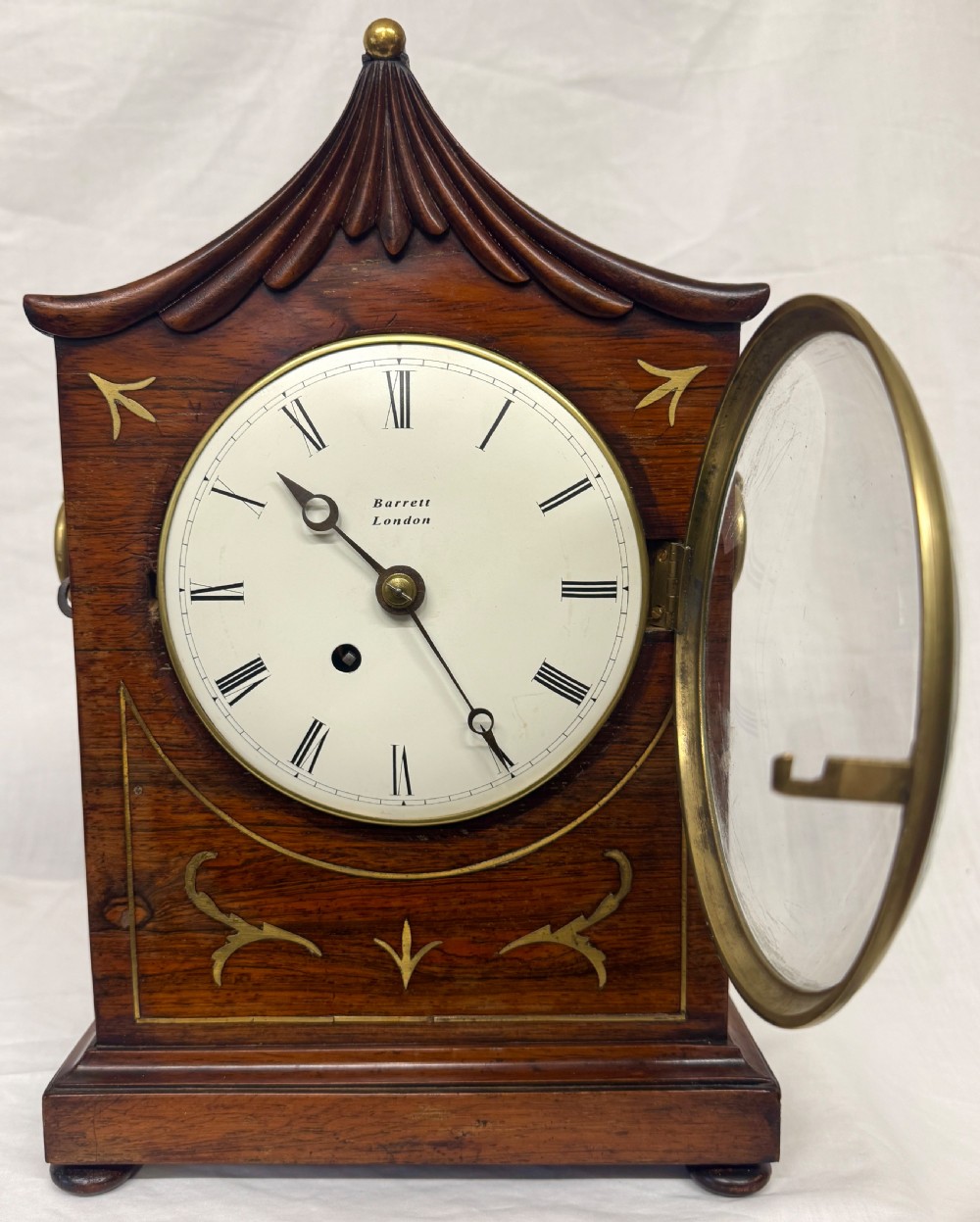 An antique mahogany mantel clock featuring a carved pagoda top, brass inlay details, side ring handles, and a white circular dial signed Barrett London with Roman numerals. View IV