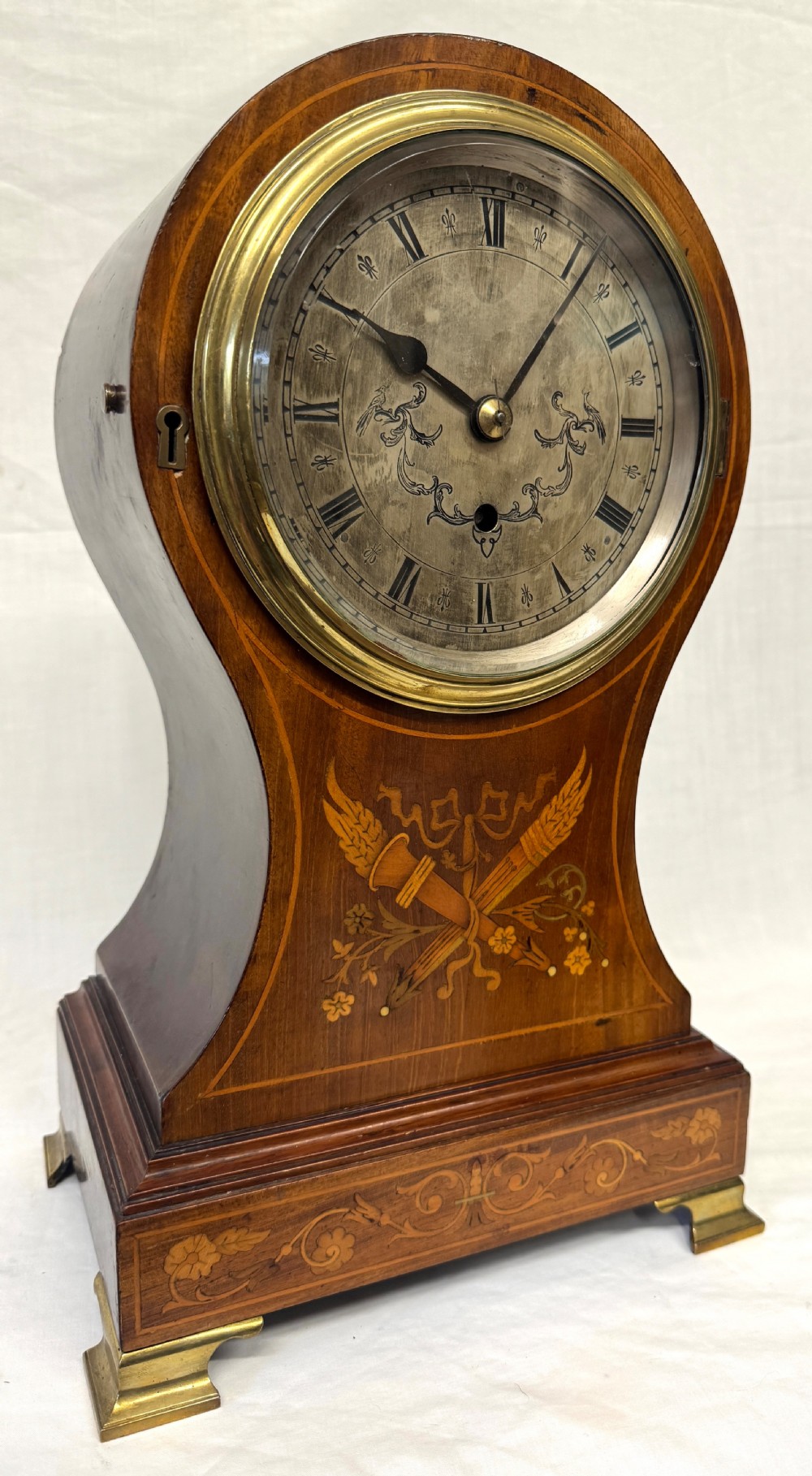 An antique balloon-shaped mahogany mantel clock featuring marquetry inlay, a silvered dial with Roman numerals, and brass bracket feet.