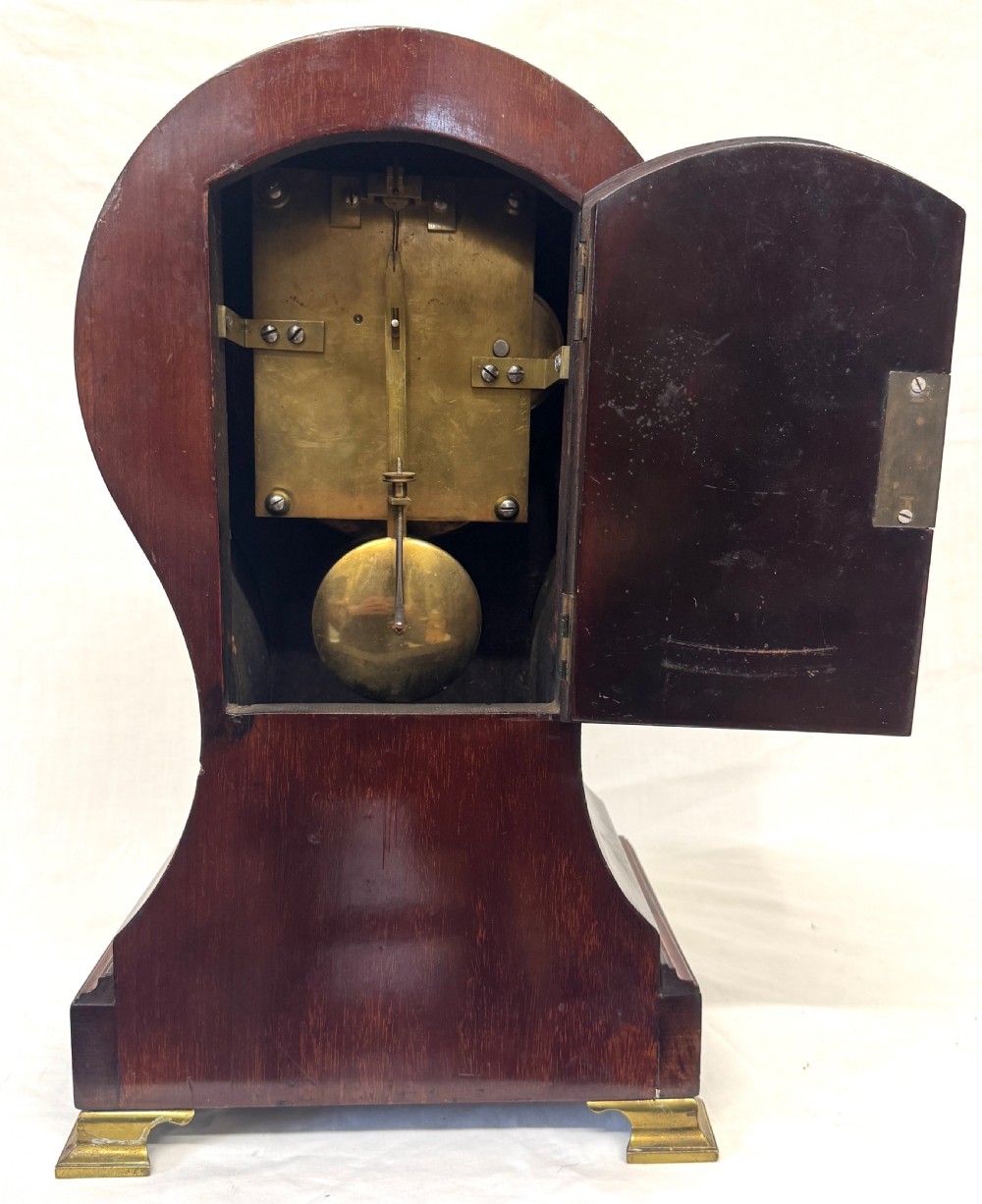 An antique balloon-shaped mahogany mantel clock featuring marquetry inlay, a silvered dial with Roman numerals, and brass bracket feet. View IX