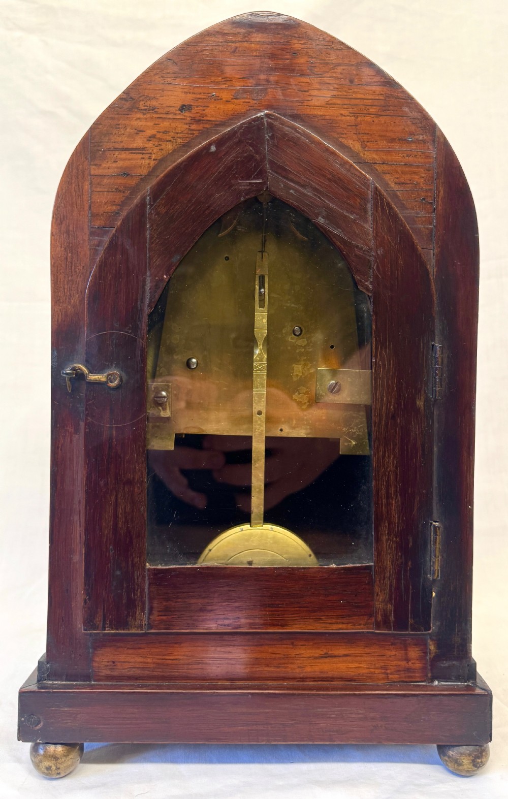 A dark mahogany lancet top mantel clock featuring brass inlay motifs, a circular white dial with Roman numerals, side ring handles, and brass ball feet. View VII