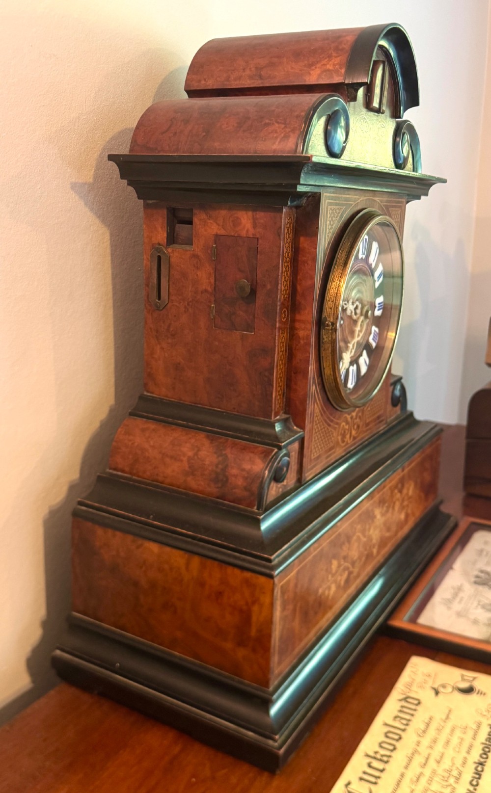 An antique walnut and ebonised cuckoo mantel clock featuring floral marquetry inlays, an architectural domed pediment, and an enamel cartouche dial. View VII