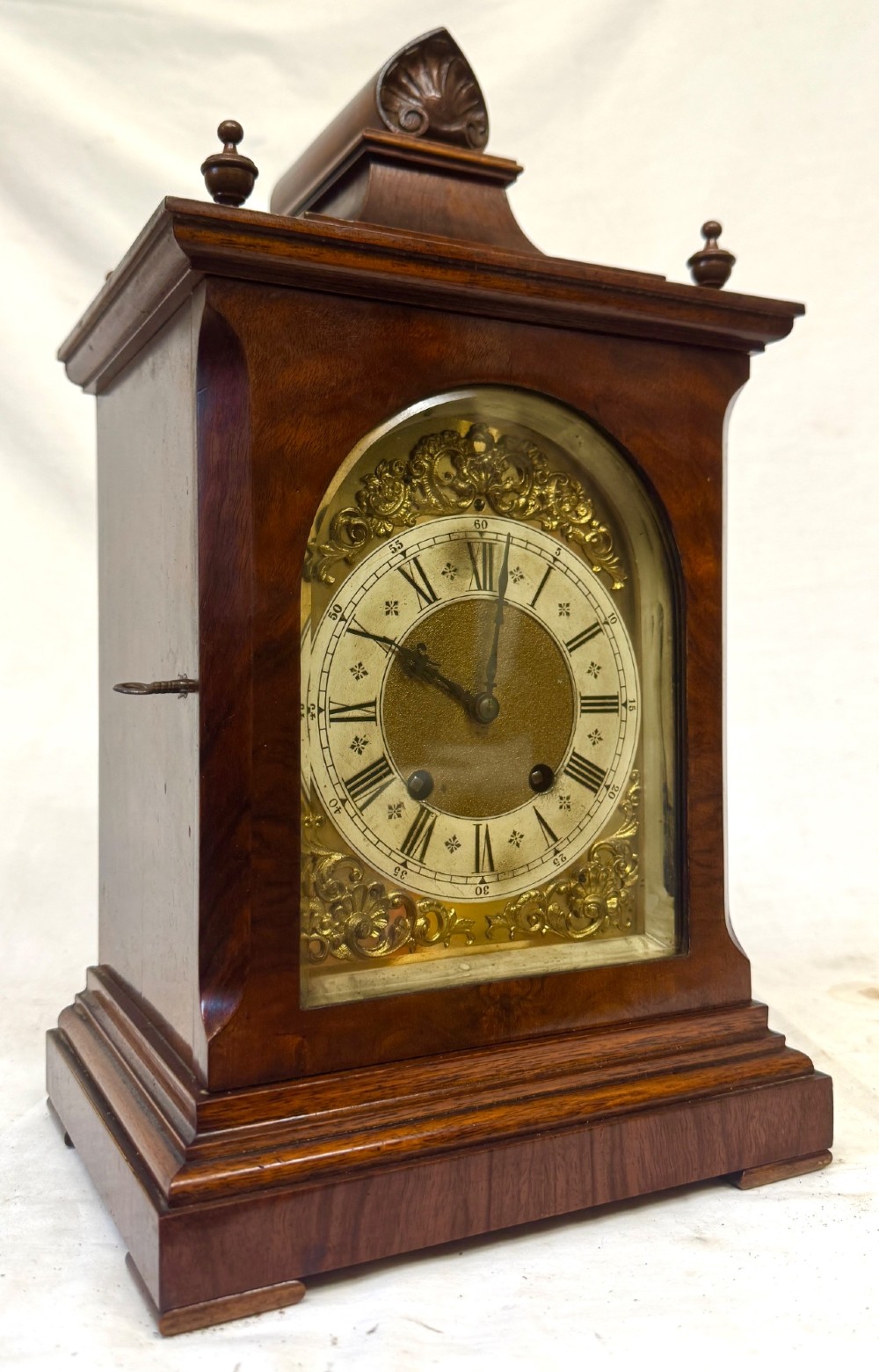 An antique walnut bracket clock featuring an arched brass dial with decorative corner spandrels, Roman numerals, and turned finials on a stepped wood base.
