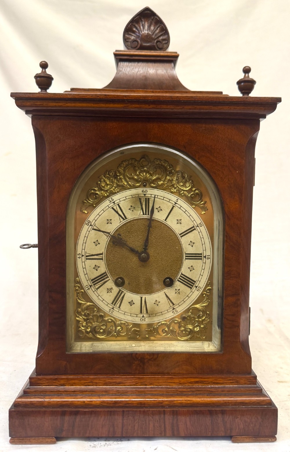 An antique walnut bracket clock featuring an arched brass dial with decorative corner spandrels, Roman numerals, and turned finials on a stepped wood base. View II